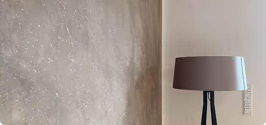 Eine graubraune Stehlampe mit rundem Lampenschirm steht vor einer verputzten Wand und einer weißen Wand mit Lichtschaltern.