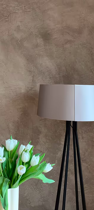 Stehlampe mit weißem Lampenschirm neben einem weißen Vase mit weißen Tulpen vor brauner Wand.