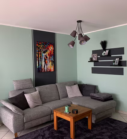 Gemütliches Wohnzimmer mit grauem Ecksofa, Holztisch und buntem Bild an der Wand.