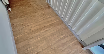 Holzboden in einem Raum mit einer weißen Wand auf der linken Seite und weißen vertikalen Jalousien auf der rechten Seite.