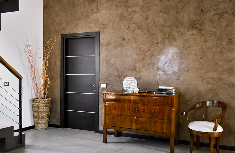 Moderner Innenraum mit schwarzer Tür, brauner Kommode, dekorativer Vase, Stuhl mit weißem Sitzkissen und Treppe mit Geländer.