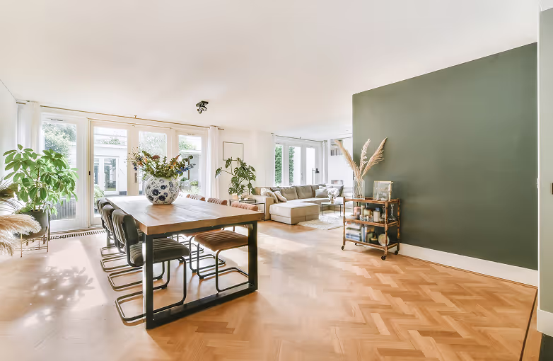 Helles Wohnzimmer mit Eichenparkett, großem Esstisch mit schwarzen Stühlen, vielen Pflanzen und einem grünen Akzentwand.