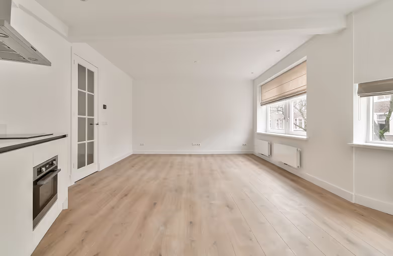 Leerer moderner Wohnraum mit hellem Holzboden, weißen Wänden, zwei großen Fenstern mit hellen Jalousien und Einbauküche mit Ofen links.
