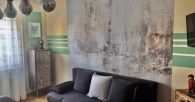 Wohnzimmer mit dunklem Sofa, einem hellen Kissen, einer abstrakten Wandgestaltung und einem Holzschrank neben einem Fenster mit weißen Vorhängen.