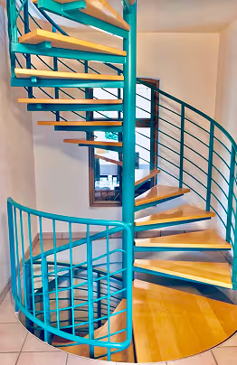 Türkisfarbene Wendeltreppe mit Holzstufen in einem hellen Innenraum neben einem Fenster.