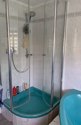 Duschkabine mit Glaswänden und grünem Boden, neben blauem Waschbecken in einem gefliesten Badezimmer.