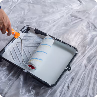 Hand hält Farbroller und taucht ihn in eine Farbwanne mit weißer Farbe auf einer durchsichtigen Plastikfolie.