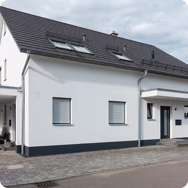 Modernes weißes Einfamilienhaus mit grauem Dach, zwei kleinen Fenstern und einer Vordertür unter einem überdachten Eingang.