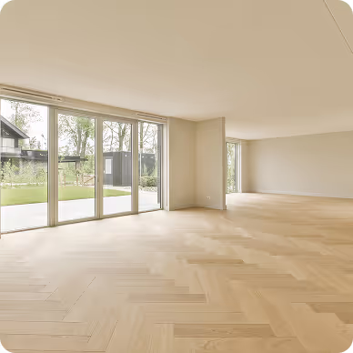 Großer, leerer Raum mit hellen Holzböden und bodentiefen Fenstern, die auf einen Garten mit Schuppen blicken.