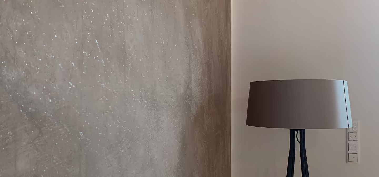 Standlampe mit beigem Lampenschirm vor einer Wand mit strukturierter grauer Oberfläche und angrenzender weißer Wand mit Lichtschaltern.