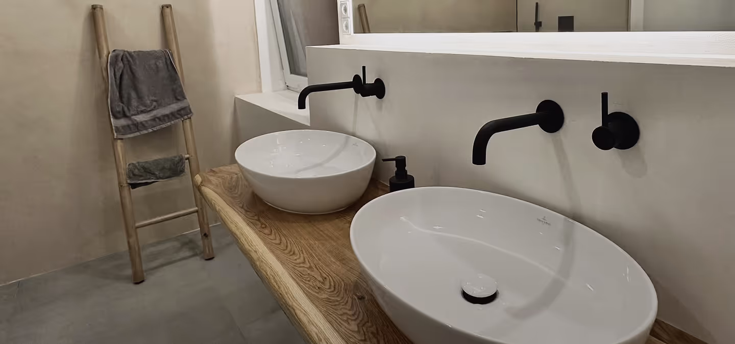 Modernes Badezimmer mit zwei weißen Waschbecken auf einer Holzplatte, schwarzen Wandarmaturen und einer hölzernen Leiter mit Handtüchern.